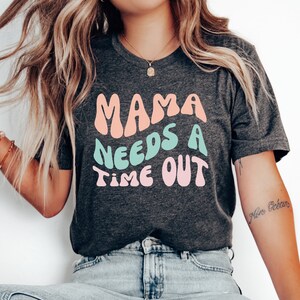 May include: A dark grey t-shirt with the text "Mama Needs a Time Out" in pink, blue, and peach colored letters.