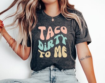 May include: A dark gray t-shirt with the text "Taco Dirty To Me" in colorful, stylized lettering.