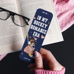 Könnte beinhalten: Marineblaue Lesezeichen mit dem Text "IN MY HOCKEY ROMANCE ERA" und einer Illustration eines Mädchens mit Kopfhörern, das ein Buch liest. Das Lesezeichen zeigt auch einen Hockeyschläger und Herzen.