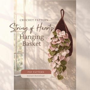 May include: A crochet pattern for a String of Hearts hanging basket. The basket is a dark brown color, with a loop for hanging. The cascading hearts are in shades of pink and green. The text on the image reads "CROCHET PATTERN String of Hearts Hanging Basket".