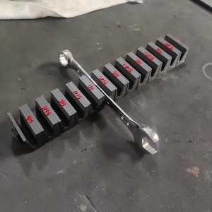 May include: A set of gray wrench organizers with red size labels. A silver wrench rests across the organizers. The organizers are on a dark surface, suggesting a workshop or garage setting. The image highlights tools and organization.