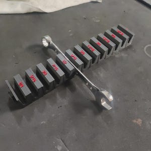 May include: A set of gray wrench organizers with red size markings. A silver wrench rests across the organizers. The sizes are in inches, including 1/4, 5/16, 3/8, 7/16, 1/2, and 9/16.
