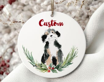 May include: A white ceramic Christmas ornament featuring a watercolour illustration of a dog, encircled by a green wreath with red berries. The word "Custom" is written in red script. A gold string is attached for hanging.