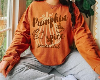 May include: A burnt orange crewneck sweatshirt featuring a large graphic of a pumpkin, an acorn, and a coffee mug. The text "Pumpkin spice social club" is printed in dark brown script and serif lettering across the center of the chest.