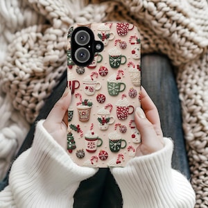 May include: A phone case featuring a festive Christmas-themed pattern. The design includes various mugs of hot chocolate, candy canes, and ornaments in red, green, and white on a light pink background. The case has a dual-camera cutout.