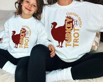May include: Two white crewneck sweatshirts featuring a dark red turkey silhouette graphic and gold text that reads CUSTOM FAMILY TURKEY TROT 2025. The design is printed on the front of each long-sleeved pullover, which are worn with black leggings.