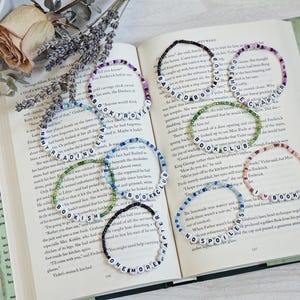 May include: A collection of beaded bracelets with white letter beads spelling out book-related words like "BOOK CLUB" and "READING ERA." The bracelets are in various colors and are displayed on an open book with dried lavender.
