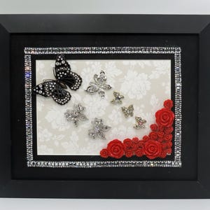 May include: A framed artwork featuring a black butterfly with white accents and several silver butterflies. Red roses adorn the lower right corner. The frame is black with a rhinestone border, set against a floral background.