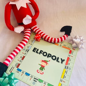 Può includere: Un gioco da tavolo a tema natalizio chiamato "Elfopoly" con un personaggio di elfo dei cartoni animati e dadi. Il tabellone è verde chiaro con spazi e illustrazioni colorate. Una bambola elfo a righe rosse e bianche è posizionata vicino al tabellone, insieme a un fiocco verde e argento.