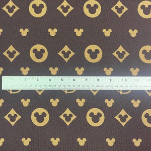 Op de afbeelding: Bruine stof met een herhaald patroon van gouden Mickey Mouse-hoofden, ruiten en het Disney-logo. Een liniaal ligt over de stof en meet tot 30 cm. De stof heeft een getextureerd uiterlijk.