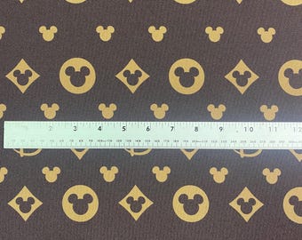 May include: Brown fabric with a repeating pattern of gold Mickey Mouse heads, diamonds, and the Disney logo. A ruler is placed across the fabric, measuring up to 12 inches. The fabric has a textured appearance.