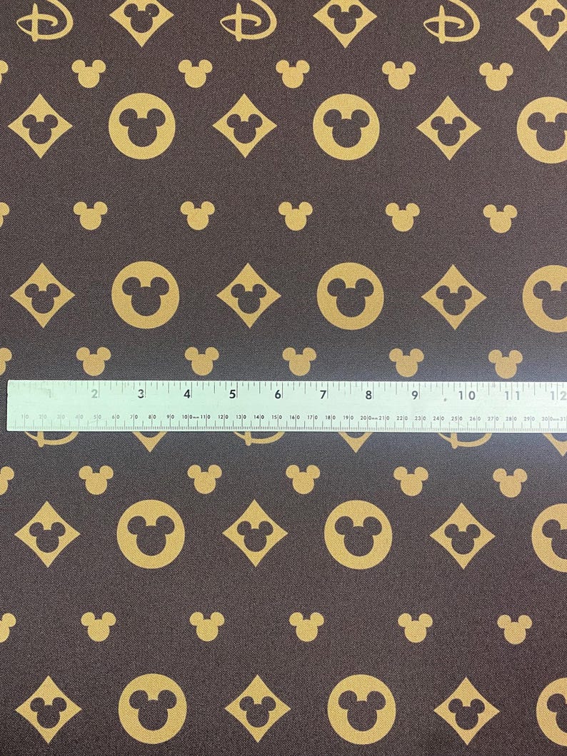 Puede incluir: Tela marr&oacute;n con un patr&oacute;n repetido de cabezas doradas de Mickey Mouse, diamantes y el logotipo de Disney. Una regla se coloca sobre la tela, midiendo hasta 30 cm. La tela tiene una apariencia texturizada.