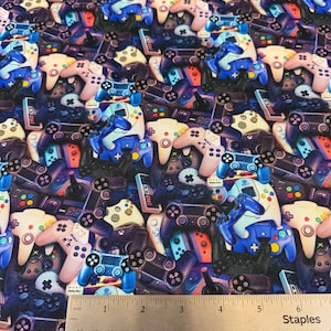 May include: Fabric with a repeating pattern of colorful video game controllers in various styles and colors, including blue, pink, and black. A ruler shows measurements in inches.