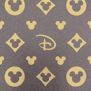 Puede incluir: Tela marr&oacute;n oscuro con un patr&oacute;n repetido de cabezas de Mickey Mouse en c&iacute;rculos y diamantes, junto con el logotipo de Disney en dorado. El fondo es de color marr&oacute;n m&aacute;s oscuro.