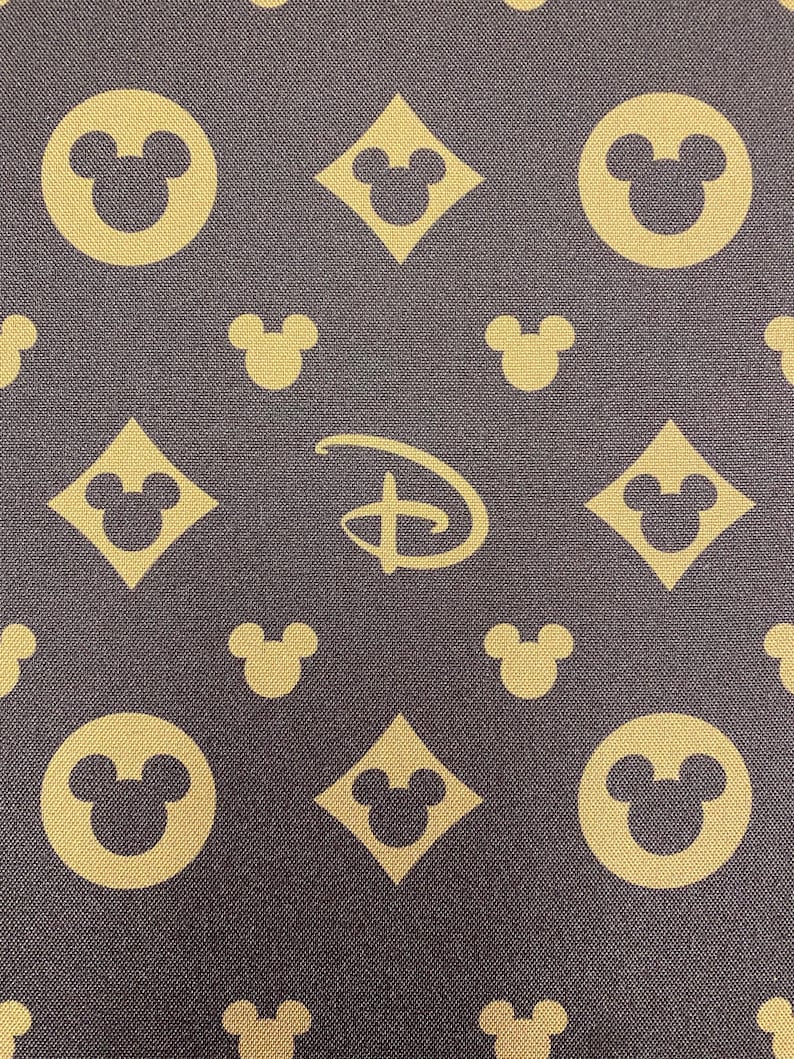 Puede incluir: Tela marr&oacute;n oscuro con un patr&oacute;n repetido de cabezas de Mickey Mouse en c&iacute;rculos y diamantes, junto con el logotipo de Disney en dorado. El fondo es de color marr&oacute;n m&aacute;s oscuro.