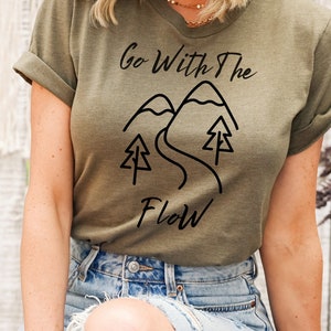 Puede incluir: Una camiseta verde con un diseño gráfico negro que dice "Go With The Flow" y presenta una cordillera con un camino sinuoso y árboles.
