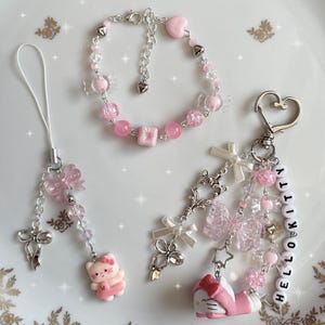 Könnte beinhalten: Eine Sammlung von rosa-silbernen Accessoires, darunter ein Armband, ein Handy-Anhänger und ein Schlüsselanhänger. Das Armband hat rosa Perlen und herzförmige Charms. Der Handy-Anhänger und der Schlüsselanhänger haben Schleifen- und Hello Kitty-Charms. Die Accessoires liegen auf einer weißen Oberfläche.