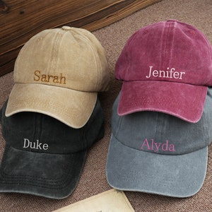 Könnte beinhalten: Vier personalisierte Baseballkappen in verschiedenen Farben. Die Kappen sind beige, burgunderrot, schwarz und grau. Die Namen "Sarah", "Jenifer", "Duke" und "Alyda" sind auf der Vorderseite der Kappen gestickt.