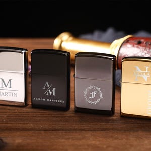 May include: Four personalized lighters in silver, black, and gold. Each lighter features custom engraved initials, names, or a monogram. The gold lighter has the word "GROOM" and a date. The lighters are on a wooden surface.