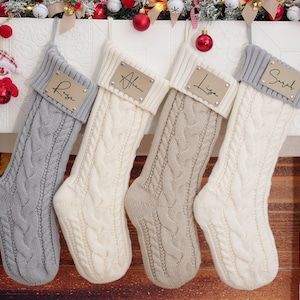 Könnte beinhalten: Vier weiße und graue gestrickte Weihnachtsstrümpfe mit personalisierten goldenen Namensschildern. Die Namen auf den Schildern sind Rose, Alan, Lisa und Sarah.