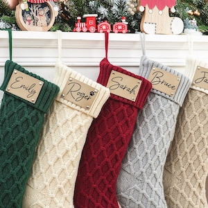 Könnte beinhalten: Fünf personalisierte Weihnachtsstrümpfe mit den Namen "Emily", "Roger", "Sarah", "Bruce" und "Janice" auf goldenen Etiketten. Die Strümpfe sind aus gestricktem Stoff in Grün, Creme, Rot, Grau und Beige.