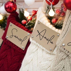 Könnte beinhalten: Vier Weihnachtsstrümpfe mit personalisierten Namensschildern. Die Strümpfe sind aus gestricktem Stoff in den Farben Grün, Rot, Weiß und Creme. Die Namensschilder sind aus braunem Leder mit weißen Buchstaben. Die Namen auf den Schildern sind Kevin, Samuel, Amelia und Justin.