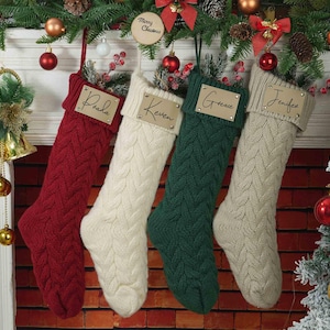 Könnte beinhalten: Vier gestrickte Weihnachtsstrümpfe in Rot, Weiß, Grün und Creme mit personalisierten Namensschildern. Die Schilder sind aus braunem Papier mit goldenen Buchstaben. Die Strümpfe hängen an einem Kaminsims aus Ziegeln.