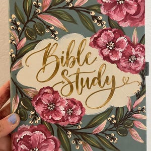 May include: A rectangular book with a sage green cover featuring hand-painted pink and dark red flowers, green leaves, and gold script text that reads Bible Study. The floral design forms a wreath around the central gold lettering on the cover.