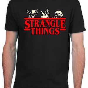 Puede incluir: Camiseta negra con un gráfico blanco de cuatro figuras en una pose de lucha. El texto "Strangle Things" está impreso en letras rojas.