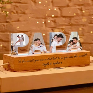 May include: A wooden light-up sign spelling "PAPA" with family photos inside each letter. The base has engraved text: "To the world you are a dad but to us you are the world! Layla & Sophia." The sign is lit with a soft blue glow.
