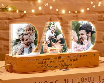 Könnte beinhalten: Ein beleuchtetes Holzschild, das "DAD" buchstabiert, mit einem Fotorahmen in jedem Buchstaben. Die Rahmen sind mit Fotos eines Vaters und seiner Kinder gefüllt. Die Basis des Schildes trägt die Inschrift "I love you more than all the stars in the sky and all the sand on the beach Mom Alayna".