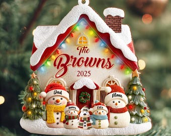 May include: A festive Christmas house ornament with the text "The Browns 2025". The ornament has a red roof, snow-covered details, and a string of lights. It features snowmen with names and two small Christmas trees.