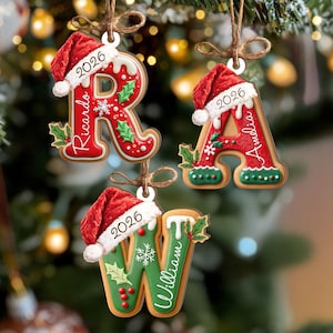 May include: Three letter-shaped Christmas ornaments shaped like gingerbread cookies. The letters R, A, and W are decorated with white icing, holly leaves, and red berries. Each letter wears a red Santa hat with the year 2026 and a name printed on it.