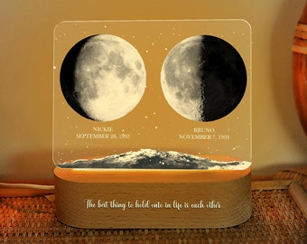 May include: A personalized night light featuring two moon phase images with dates and names. The light has a wooden base and a clear acrylic panel with the text "The best thing to hold onto in life is each other."