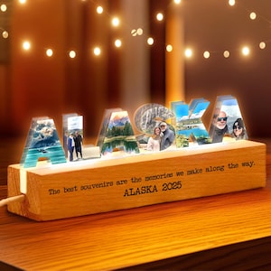 May include: A wooden light-up sign spelling "ALASKA" with photos inside each letter. The base of the sign has the text "The best souvenirs are the memories we make along the way. ALASKA 2025". Warm lights are strung above.