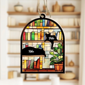 May include: A stained glass ornament featuring a bookshelf with two cats, one named "Milo" and the other named "Niko". The ornament is decorated with colorful books and a green plant.