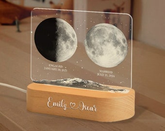 May include: A wooden base with a personalized inscription "Emily Oscar" and a heart symbol. The base holds a clear acrylic panel with two moon phases, one depicting the moon on January 18, 2021, labeled "Engaged", and the other depicting the moon on July 31, 2021, labeled "Married".