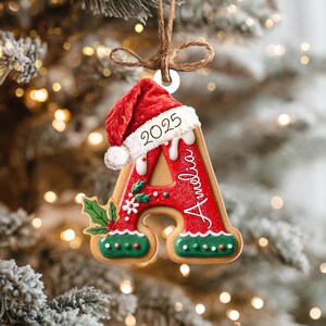 Pu&ograve; includere: Un ornamento natalizio a forma di lettera "A" con un cappello da Babbo Natale. L'ornamento rosso presenta l'anno "2025" e il nome "Amelia". Decorato con agrifoglio e glassa verde, &egrave; appeso a un anello di spago.