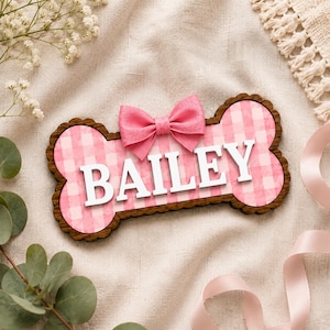 Puede incluir: Un letrero de madera con forma de hueso que presenta un fondo de patr&oacute;n de cuadros vichy rosa y blanco con el nombre BAILEY en letras may&uacute;sculas blancas en relieve. Un lazo de tela rosa est&aacute; unido al centro superior del letrero, con borde festoneado.