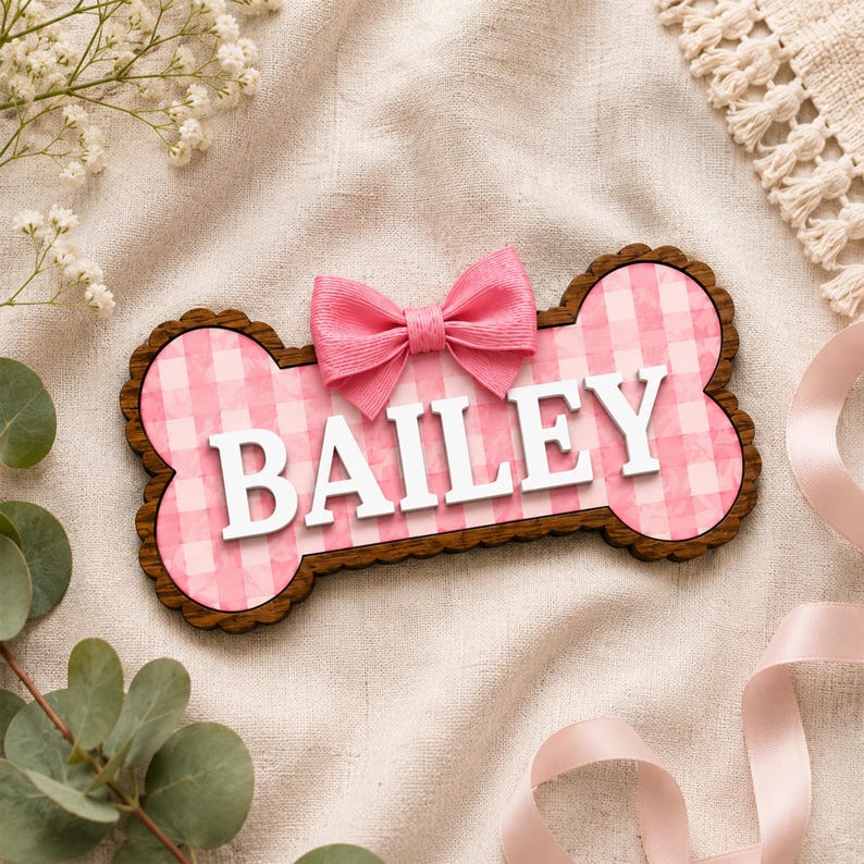 Puede incluir: Un letrero de madera con forma de hueso que presenta un fondo de patr&oacute;n de cuadros vichy rosa y blanco con el nombre BAILEY en letras may&uacute;sculas blancas en relieve. Un lazo de tela rosa est&aacute; unido al centro superior del letrero, con borde festoneado.