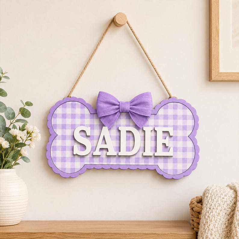 Puede incluir: Un letrero de pared con forma de hueso que presenta un patr&oacute;n de cuadros vichy en color lila claro y blanco. El letrero muestra el nombre SADIE en letras may&uacute;sculas blancas en relieve en el centro. Un lazo de tela lila claro est&aacute; sujeto sobre el nombre y una cuerda de yute est&aacute; fijada en la parte superior para colgarlo.