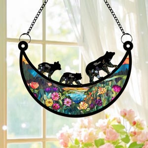 May include: A stained glass moon-shaped suncatcher with a floral design and three black bear silhouettes. The bears are walking across the moon and each bear has a name: Mom, Mia, and Ben.