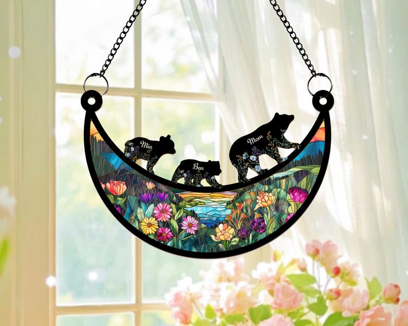 May include: A stained glass moon-shaped suncatcher with a floral design and three black bear silhouettes. The bears are walking across the moon and each bear has a name: Mom, Mia, and Ben.