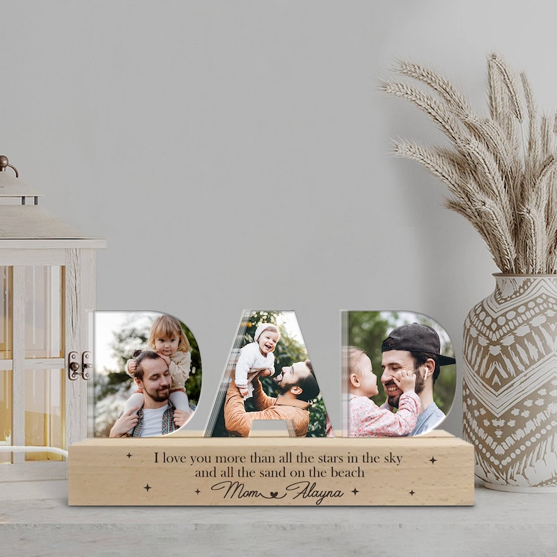 May include: A wooden photo frame shaped like the word "DAD" with three separate photo slots. Each slot features a photo of a father and child. The base of the frame has a woodgrain finish and includes the text "I love you more than all the stars in the sky and all the sand on the beach + Mom Alayna +"