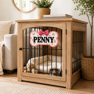 Puede incluir: Una jaula de madera para mascotas con barrotes de metal negro y acabado en madera clara. En el frente, un letrero en forma de hueso con estampado floral y un lazo rosa muestra el nombre PENNY en letras negras. Dentro hay una cama blanca y un cuenco.