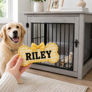 Puede incluir: Un letrero con forma de hueso con un patr&oacute;n de cuadros vichy amarillo y blanco y un lazo amarillo, con el nombre RILEY en letras negras en negrita. El letrero se sostiene frente a una jaula de madera gris con barrotes negros y una cama gris.