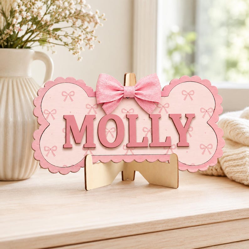 Puede incluir: Un letrero de madera con forma de hueso y fondo rosa claro con un patr&oacute;n repetitivo de peque&ntilde;os lazos. El nombre MOLLY aparece en letras may&uacute;sculas grandes de color rosa oscuro en el centro. Un lazo de tela rosa texturizada est&aacute; fijado arriba.