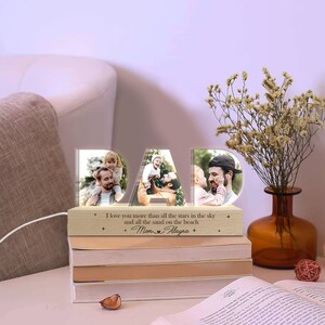 May include: A wooden photo frame shaped like the word "DAD" with a light inside. The frame has three photos of a father and child. The base of the frame has the text "I love you more than all the stars in the sky and all the sand on the beach. Mom Alayna".