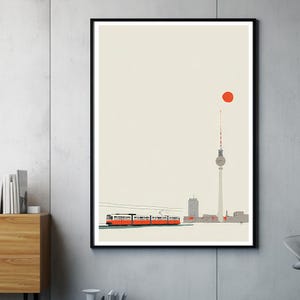 May include: A framed art print depicting a minimalist illustration of Berlin. A red tram travels along a track in front of the city's skyline, including the TV Tower. A red sun is in the sky above the tower. The artwork is in a black frame.