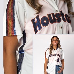 Può includere: Maglia da baseball bianca con la scritta "Houston" in paillettes blu navy e arancioni. La maglia ha un colletto blu navy e una striscia di paillettes sulle maniche. La maglia è abbottonata.